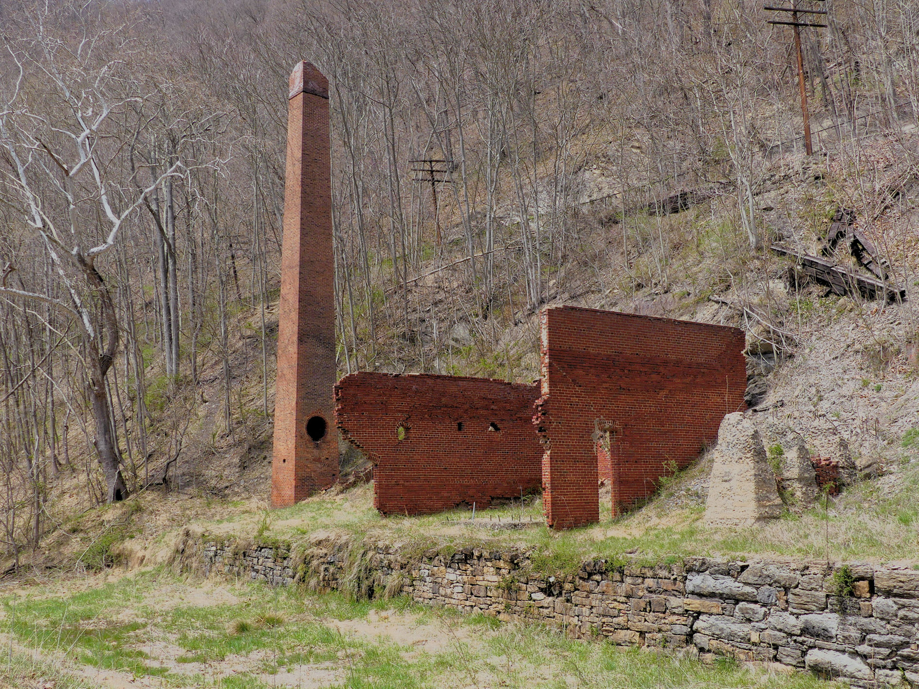 3/4 view of 19th century cement mill ruins. Furnace tower and two structural walls atop a walled embankment at the bottom of steep hill below railroad tracks.