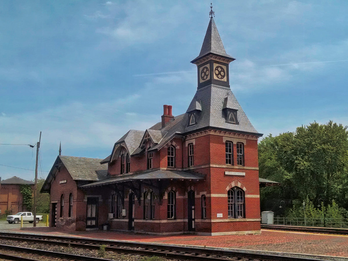 3/4 view of 19th century train station on a clear blue day. Red Brick construction with a steeple.