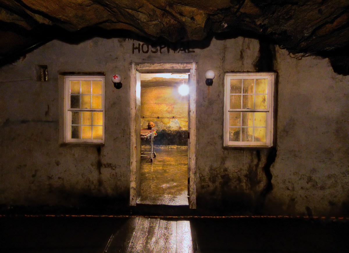 facade of a makeshift hospital inside of a coal mine. Light is shining through the doorway and windows into the darkness of the foreground.